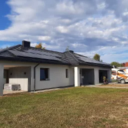 Nowoczesny dom jednorodzinny z ciemną dachówką i panelami słonecznymi na dachu, na trawniku zaparkowany biały samochód dostawczy z napisem FOTOWOLTAIKA, błękitne niebo z białymi chmurami w tle.