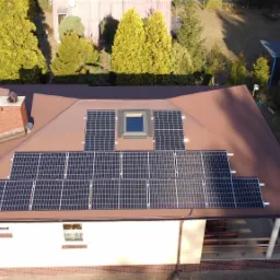 Widok z lotu ptaka na brązowy dach domu jednorodzinnego z zamontowanymi panelami słonecznymi i oknem dachowym, w tle zielone drzewa i zaparkowany samochód.