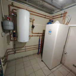 Instalacja kotłowni: biały zasobnik ciepłej wody Galmet, szary zbiornik wyrównawczy, pionowa biała pompa ciepła LG Therma V, orurowanie miedziane, płytki podłogowe w kotłowni.