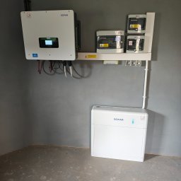 Nasz Prąd S.A. Fotowoltaika Pompy ciepła Lubliniec - Biały inwerter SOFAR, rozdzielnice i akumulator na szarej ścianie w rogu pomieszczenia. Instalacja fotowoltaiczna wewnątrz budynku.