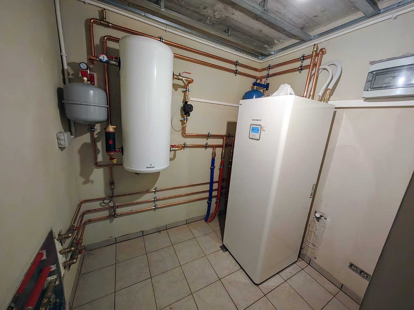 Instalacja kotłowni: biały zasobnik ciepłej wody Galmet, szary zbiornik wyrównawczy, pionowa biała pompa ciepła LG Therma V, orurowanie miedziane, płytki podłogowe w kotłowni.