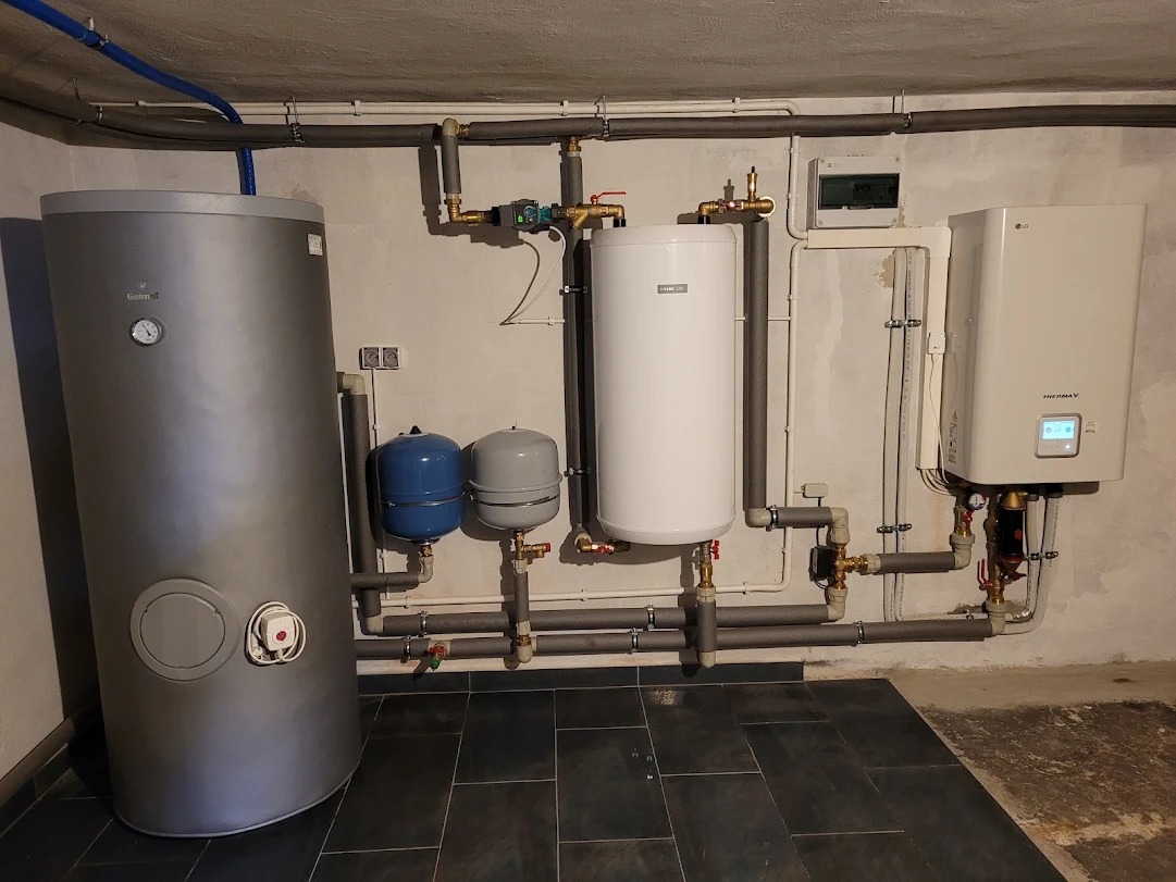 Instalacja grzewcza w kotłowni: szary zasobnik ciepłej wody, biały bojler, pompa ciepła LG Therma V z wyświetlaczem, dwa zbiorniki wyrównawcze, rury izolowane szarą pianką, płytki podłogowe...