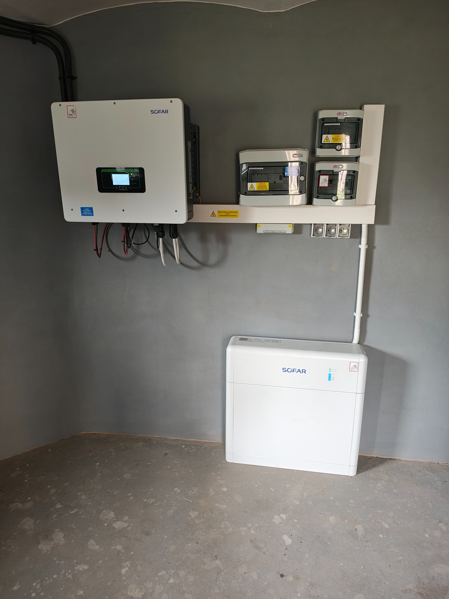 Biały inwerter SOFAR, rozdzielnice i akumulator na szarej ścianie w rogu pomieszczenia. Instalacja fotowoltaiczna wewnątrz budynku.