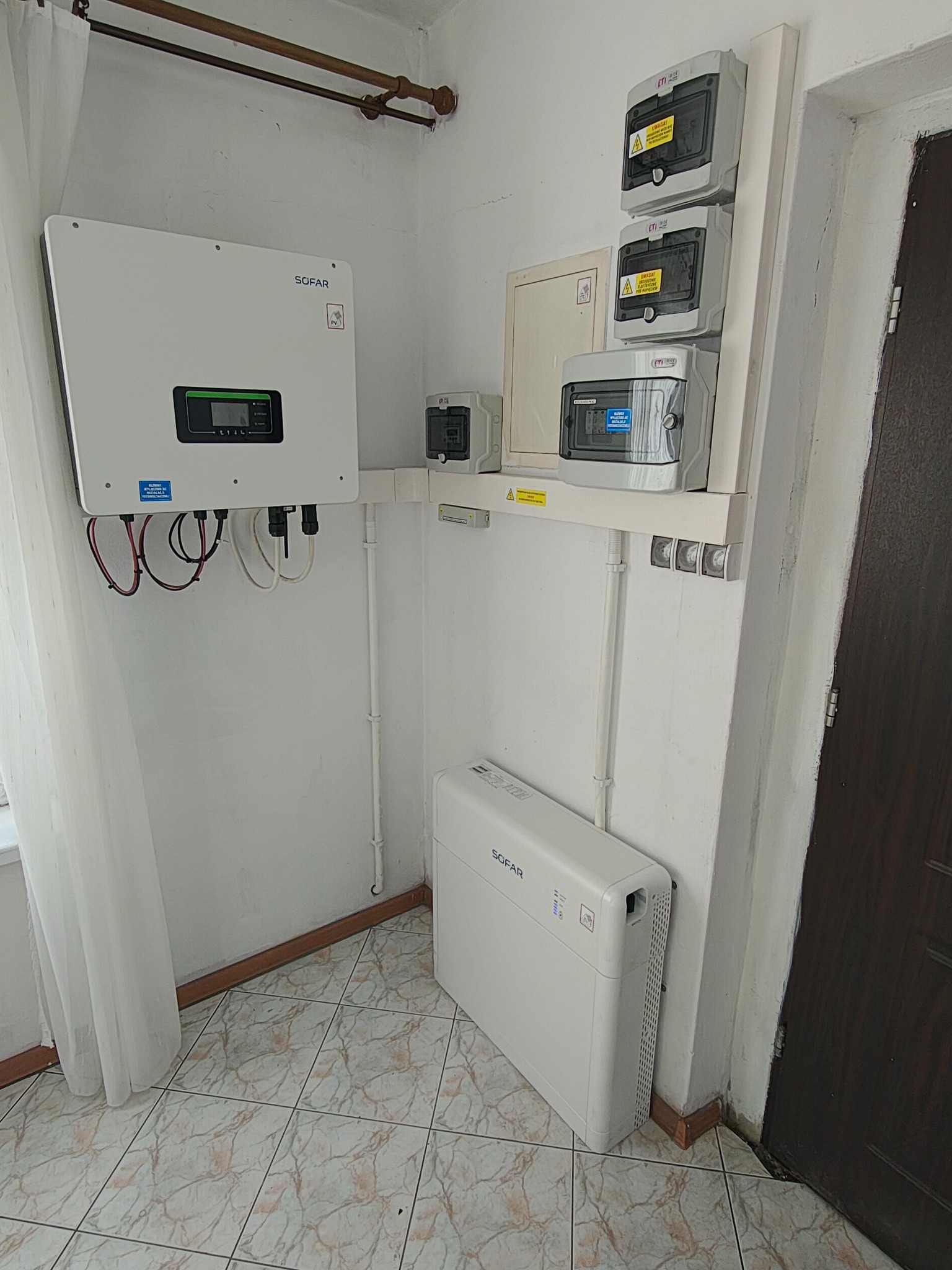 Biały falownik Sofar z podłączonymi kablami na ścianie, obok skrzynka z bezpiecznikami i biała pompa ciepła. Widoczne elementy instalacji w pomieszczeniu.