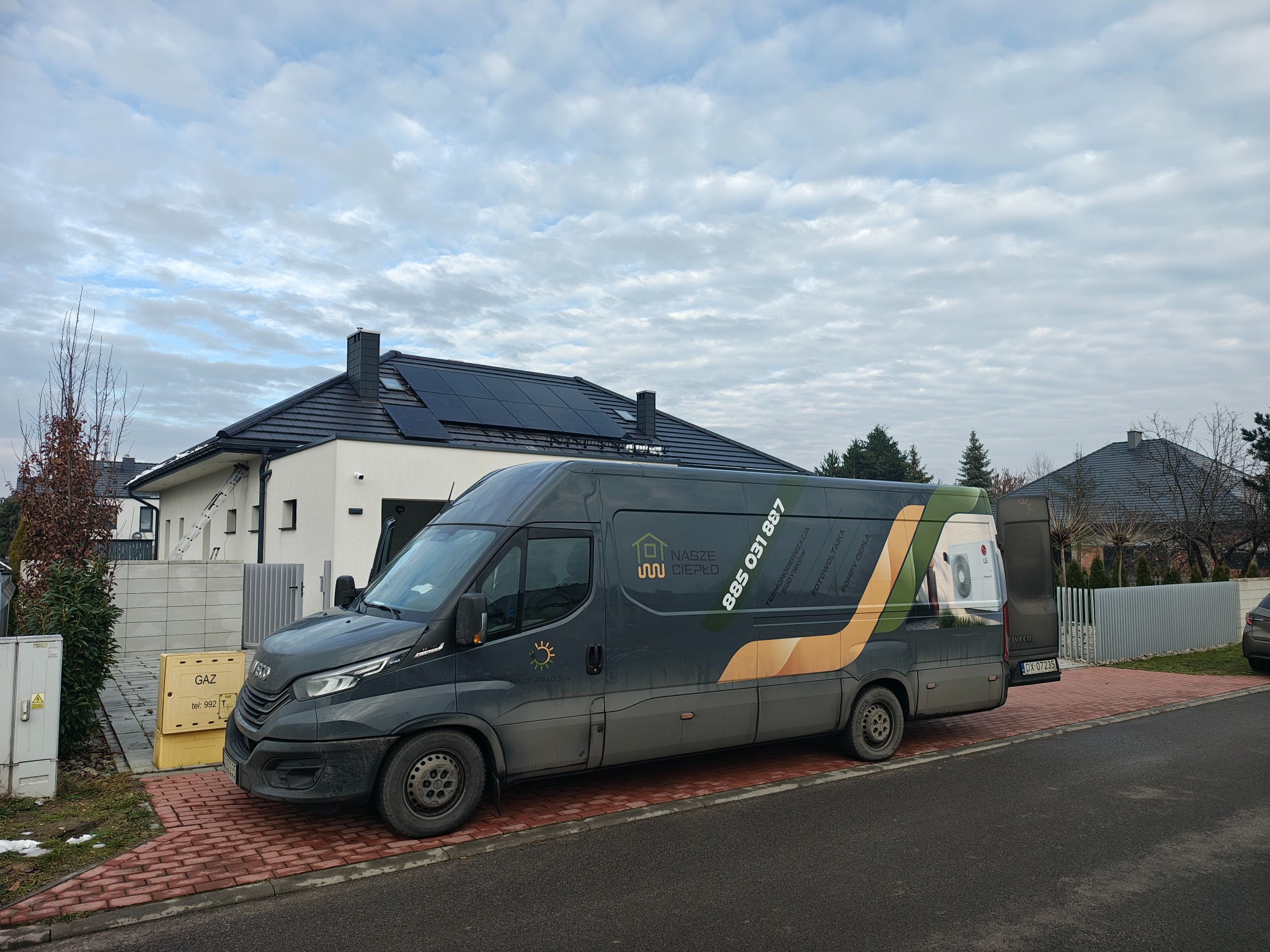 Szary furgon z logo firmy 'Nasze Ciepło' na tle nowoczesnego domu z panelami słonecznymi na dachu. Widoczny fragment pompy ciepła na tylnych drzwiach furgonu.