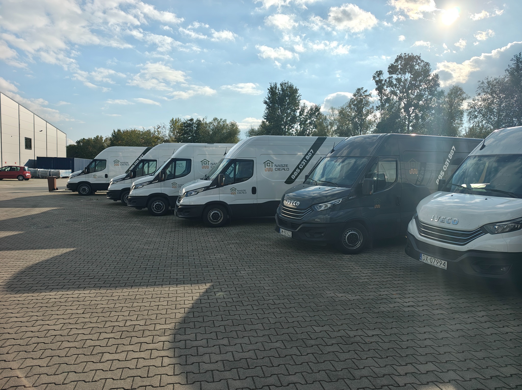 Flota firmowa: równe rzędy białych i grafitowych furgonetek Iveco z logo 'Nasze Ciepło' na tle błękitnego nieba i budynku magazynowego.