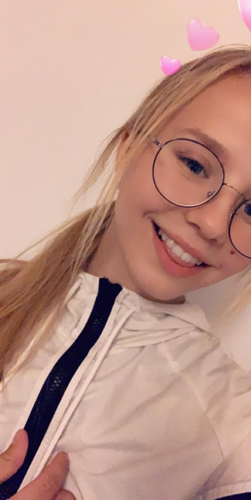 Uśmiechnięta blondynka w okularach, z filtrem serduszek, ubrana w białą bluzę z czarnym zamkiem.