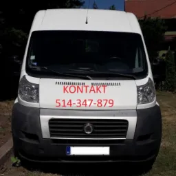 Biały bus z czerwonym napisem 'KONTAKT' i numerem telefonu na przedniej szybie, zaparkowany na trawniku przed drzewami i huśtawką.