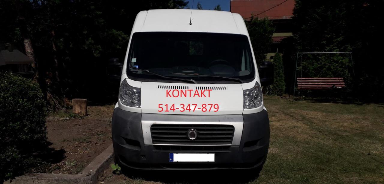 Biały bus z czerwonym napisem 'KONTAKT' i numerem telefonu na przedniej szybie, zaparkowany na trawniku przed drzewami i huśtawką.