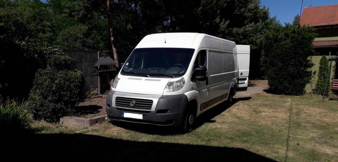 Biały bus dostawczy Fiat Ducato zaparkowany na trawie, widoczny z przodu i boku, z drugim busem w tle, w otoczeniu zieleni i ogrodzenia.