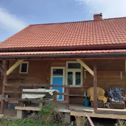 Drewniany dom z czerwonym dachem z blachy trapezowej, widoczny z zewnątrz z werandą z meblami i niebieskimi drzwiami wejściowymi.