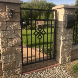 Metalowa furtka z geometrycznym wzorem, osadzona między dwoma słupkami z jasnych, teksturowanych bloczków. Za furtką widoczny trawnik i zieleń ogrodu. Po prawej stronie furtki skrzynka na listy.