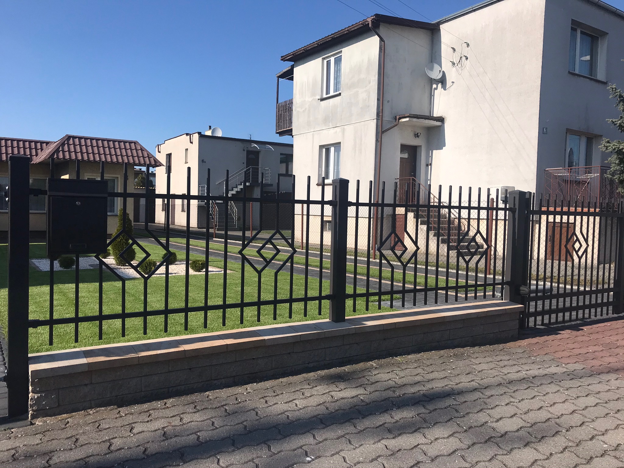 Nowoczesne ogrodzenie metalowe z geometrycznym wzorem rombu, zamontowane na niskim murku z kostki, widok na zadbany trawnik i budynki mieszkalne w tle.