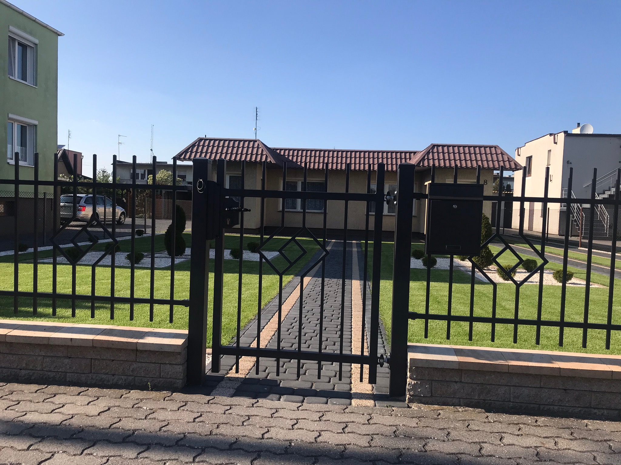 Nowoczesna, czarna, metalowa brama z geometrycznym wzorem, prowadząca do domu z kostki brukowej, otoczona trawnikiem i ozdobnymi krzewami.