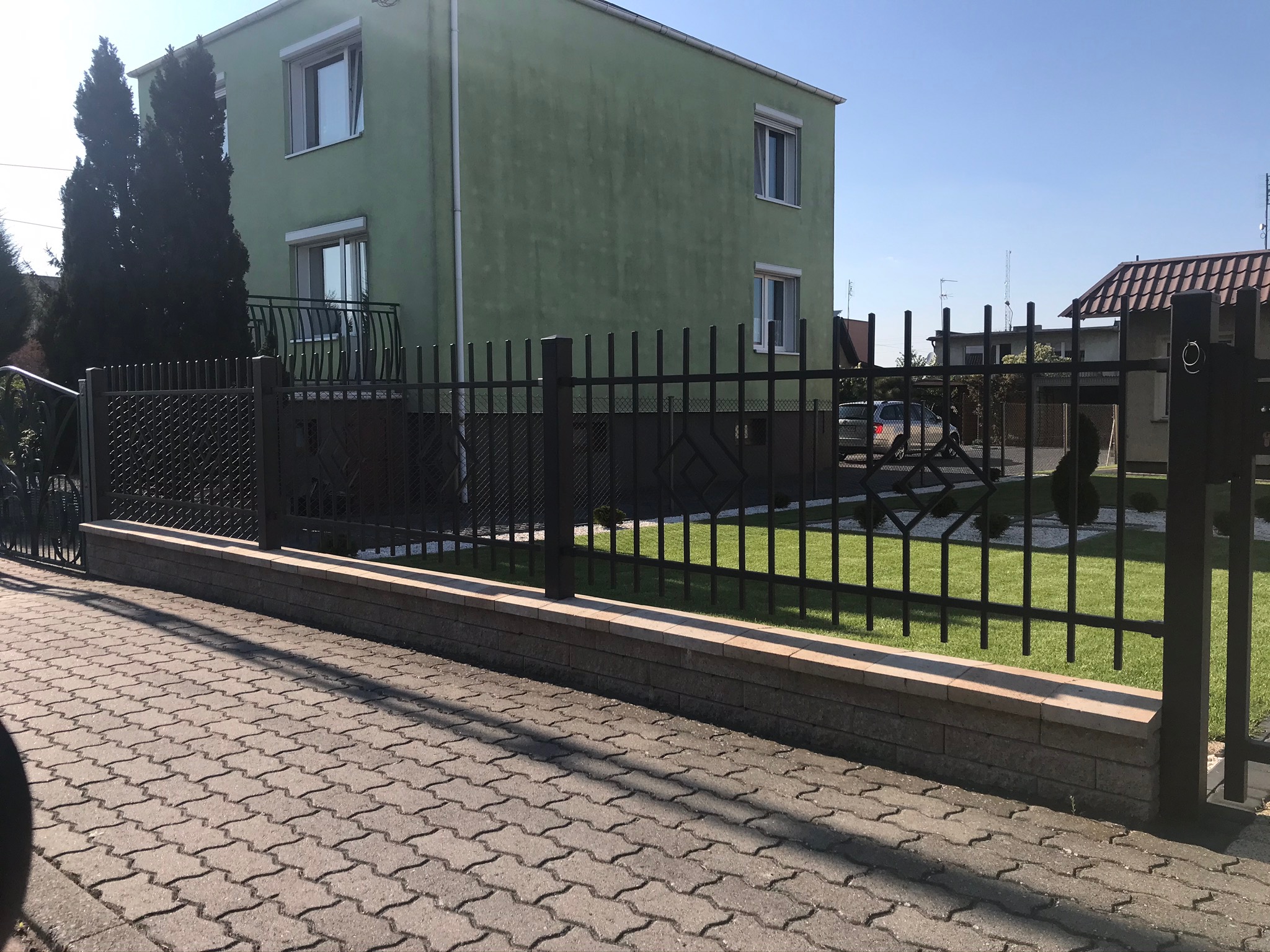 Nowoczesne, czarne ogrodzenie metalowe z elementami siatki, osadzone na niskim murku z kamienia, na tle zielonego trawnika i domu z zieloną elewacją.