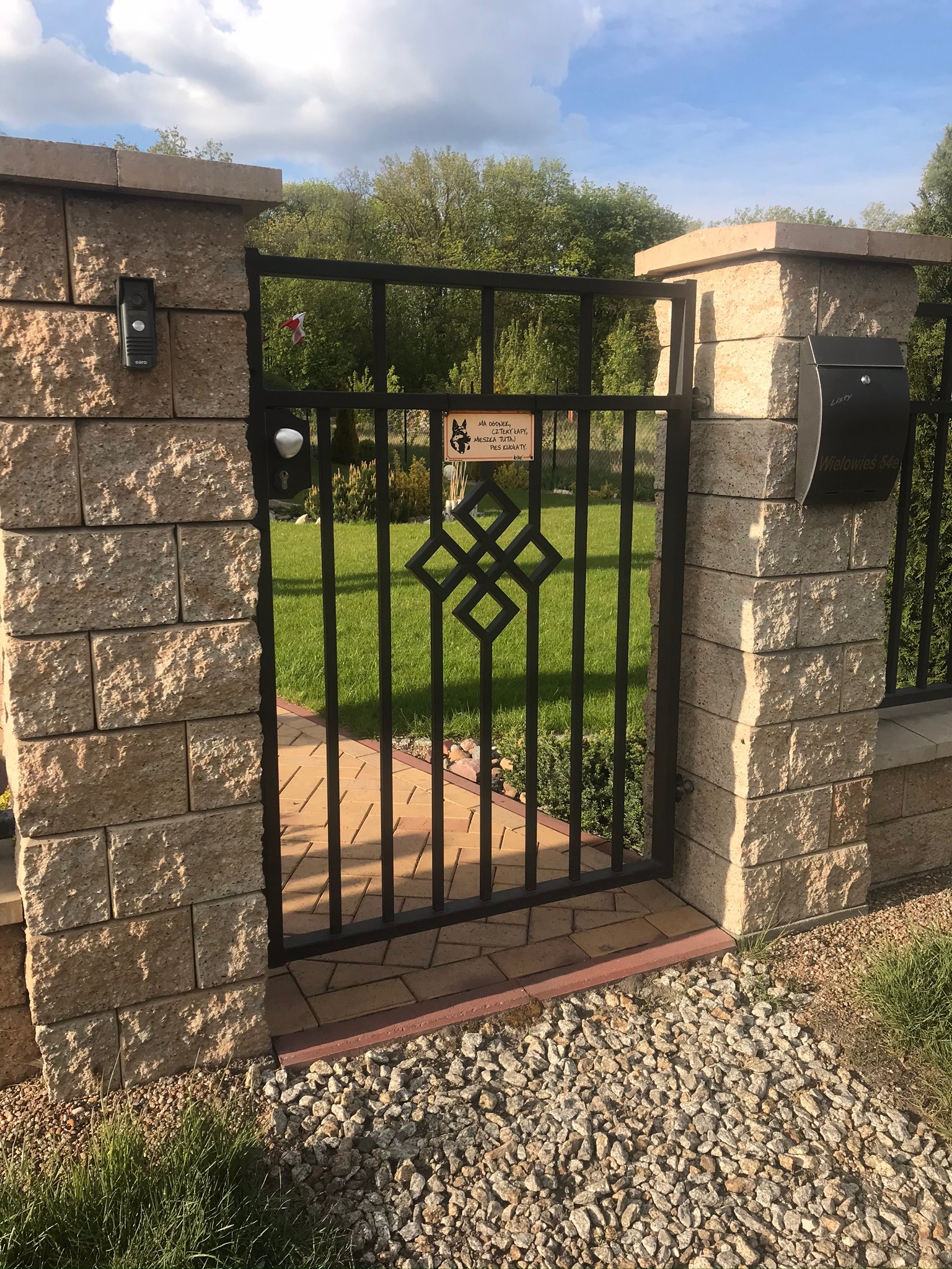 Metalowa furtka z geometrycznym wzorem, osadzona między dwoma słupkami z jasnych, teksturowanych bloczków. Za furtką widoczny trawnik i zieleń ogrodu. Po prawej stronie furtki skrzynka na listy.