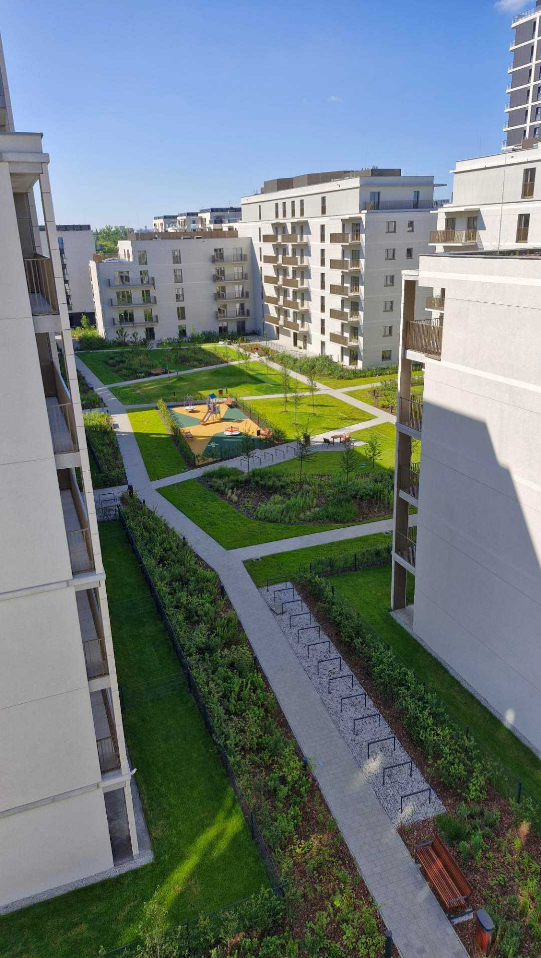 Z góry: zadbany teren zielony osiedla z placem zabaw, ścieżkami i ławkami. Nowoczesne, jasne budynki mieszkalne w tle. Regularny układ zieleni i chodników.