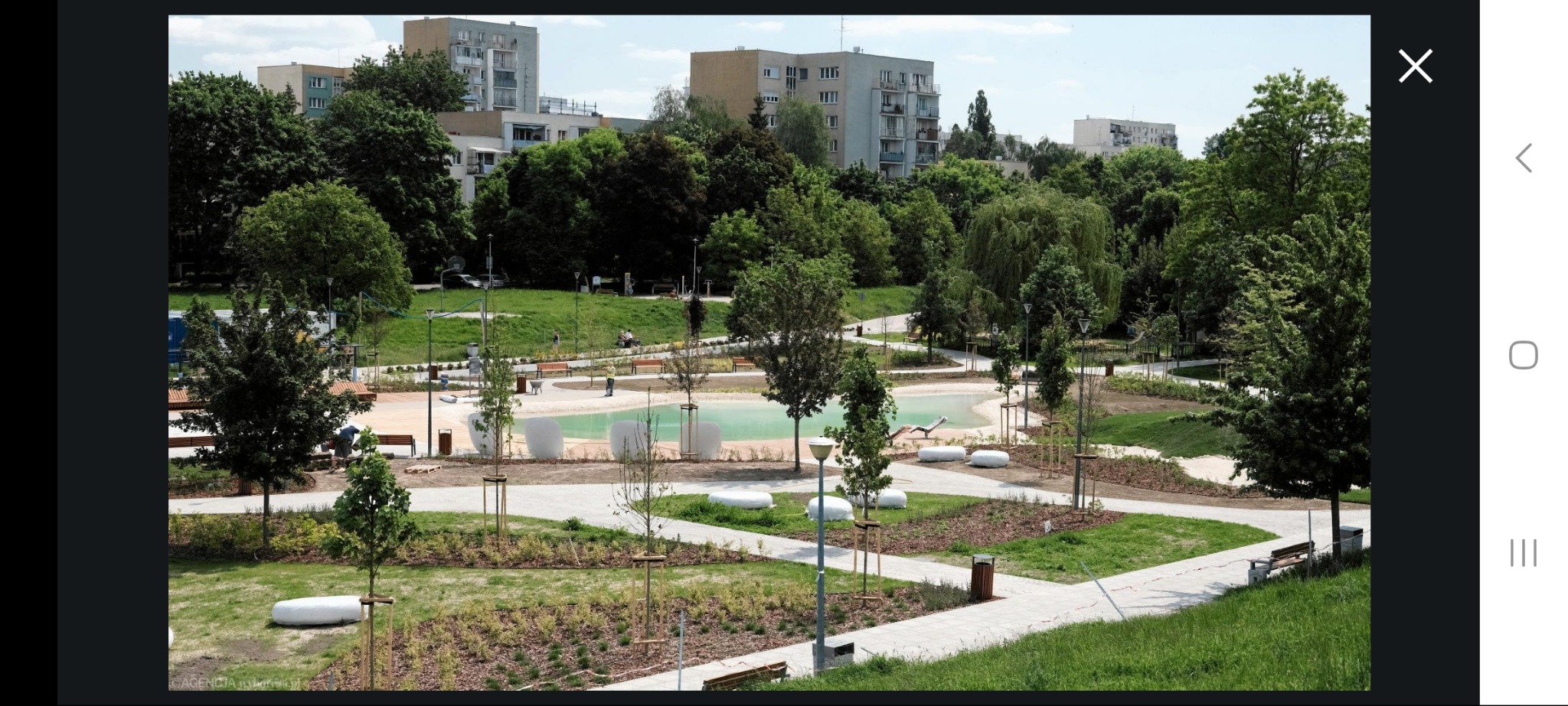 Nowo zagospodarowany park miejski z alejkami, małym stawem i nasadzeniami drzew. W tle bloki mieszkalne, a w kadrze ławki i lampy parkowe.