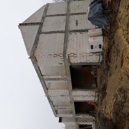 Roman Kłak - Wznoszenie dwukondygnacyjnego budynku z białych bloczków na fundamencie, widoczne zbrojenia i betonowe słupy, ziemia wokół placu budowy.