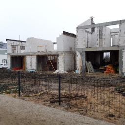 Roman Kłak - Surowy stan budowy domu z białych bloczków, widoczne niezadaszone pomieszczenia, szalunki i rusztowania, teren wokół ogrodzony siatką, w tle stojący biały samochód osobowy.