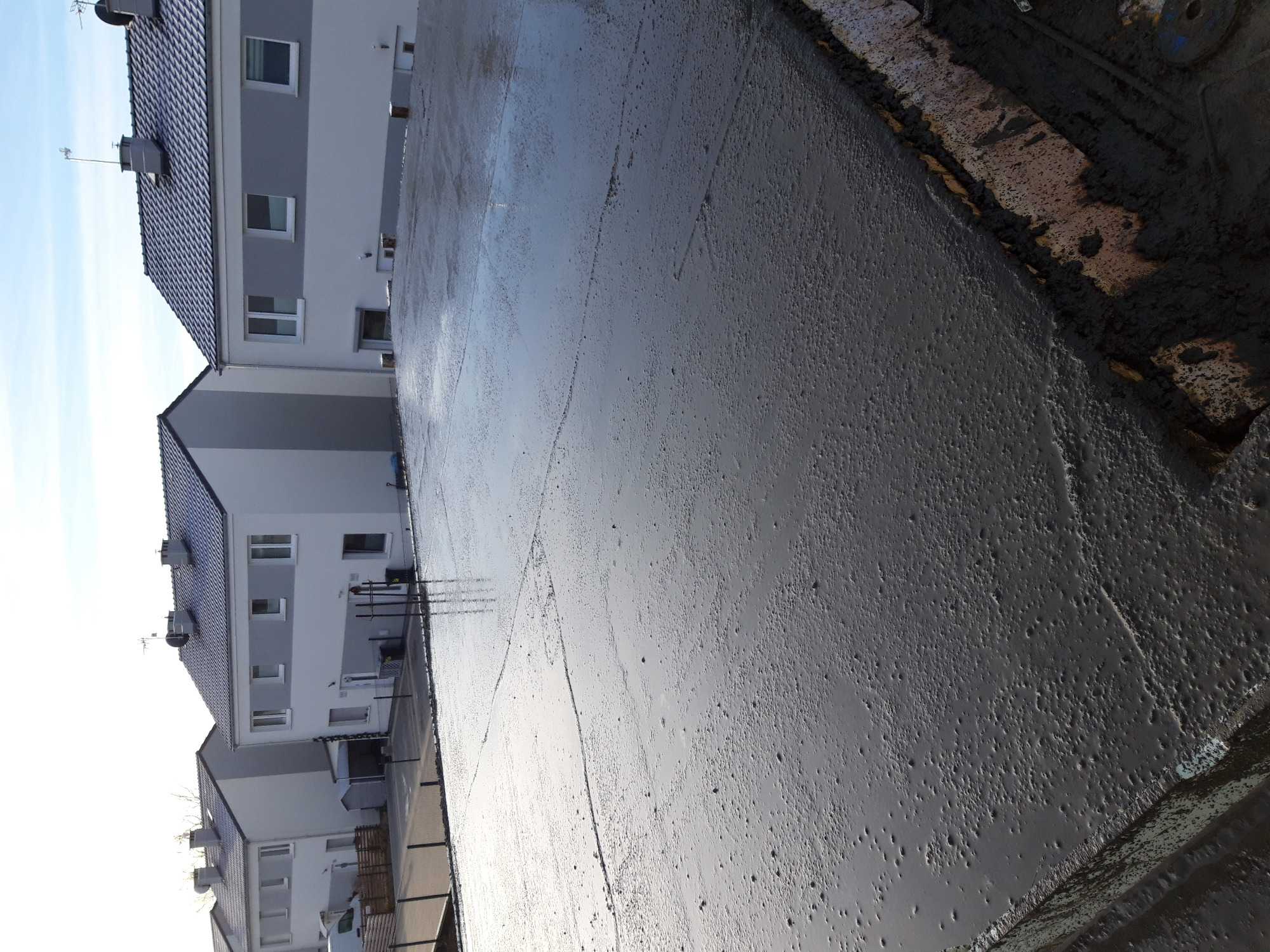 Świeżo wylany, mokry beton fundamentowy na tle budynków mieszkalnych w słoneczny dzień.