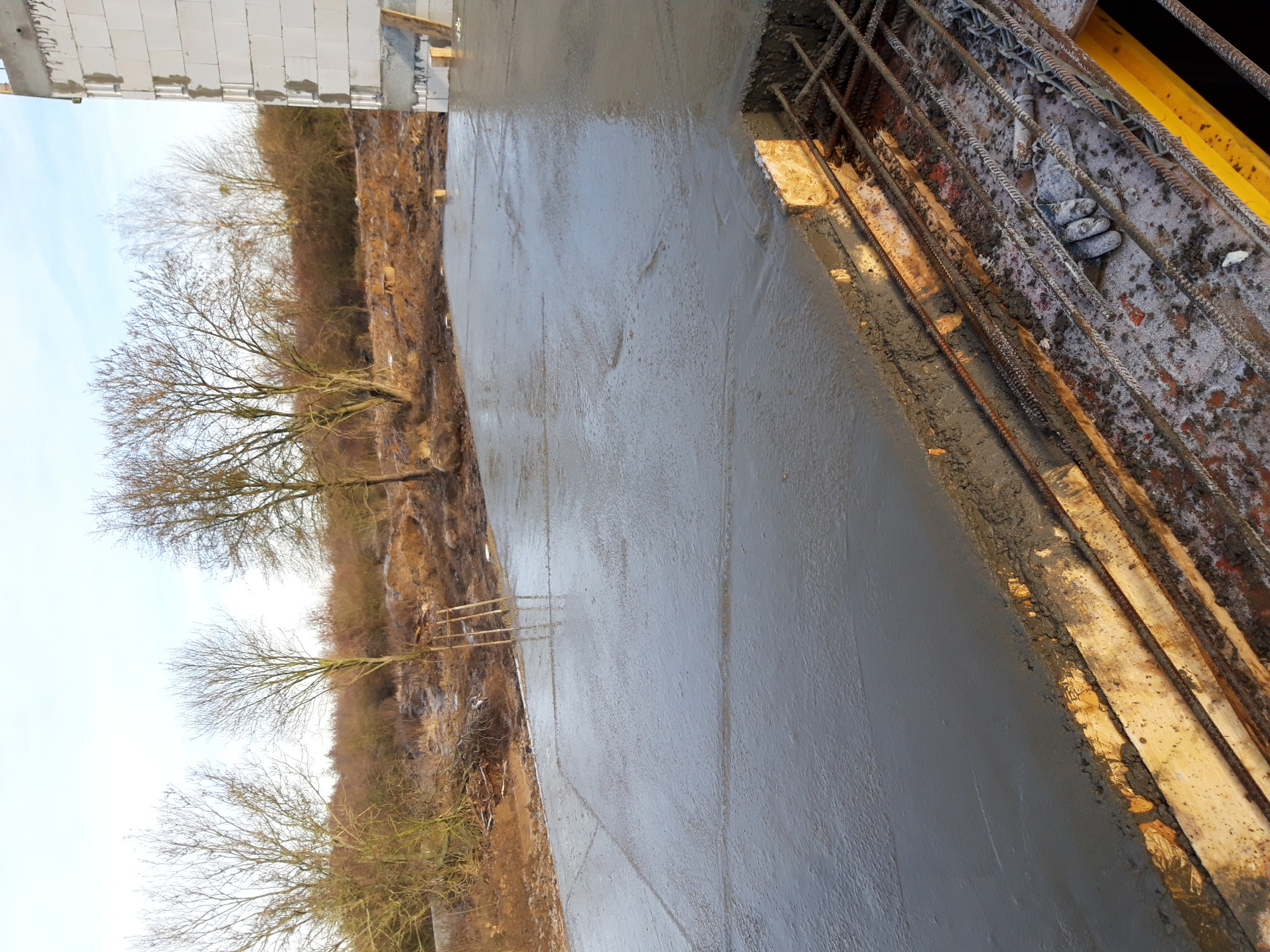 Świeżo wylany beton na przygotowany zbrojony fundament, widoczne deski szalunkowe i otaczający teren z drzewami bez liści w tle.