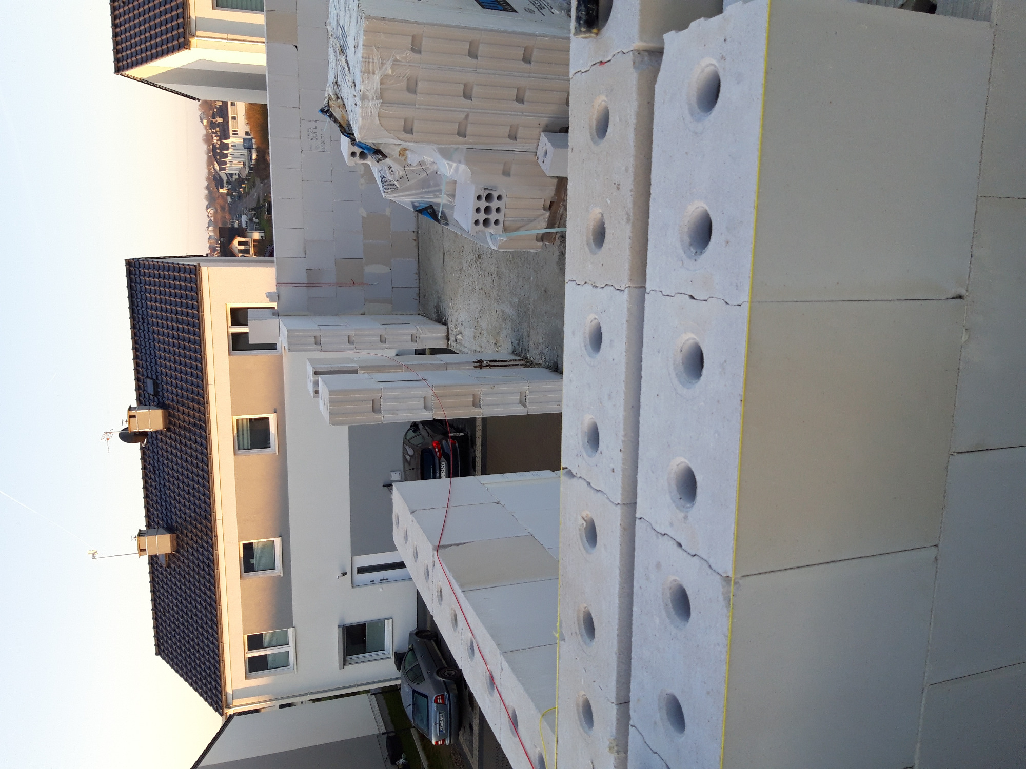 Wznoszenie ściany z betonu komórkowego Ytong na budowie domu jednorodzinnego, widoczne pakiety bloczków i fragmenty sąsiednich budynków.