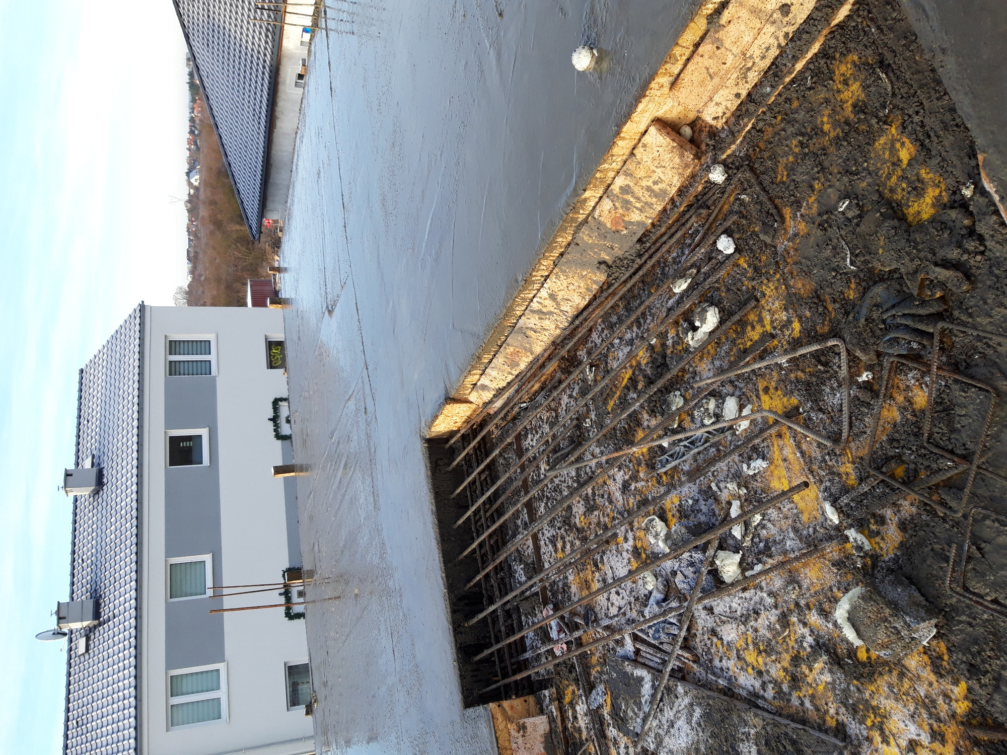 Zalewanie betonem zbrojenia fundamentu domu, widoczne szalunki i fragment budynku w tle.