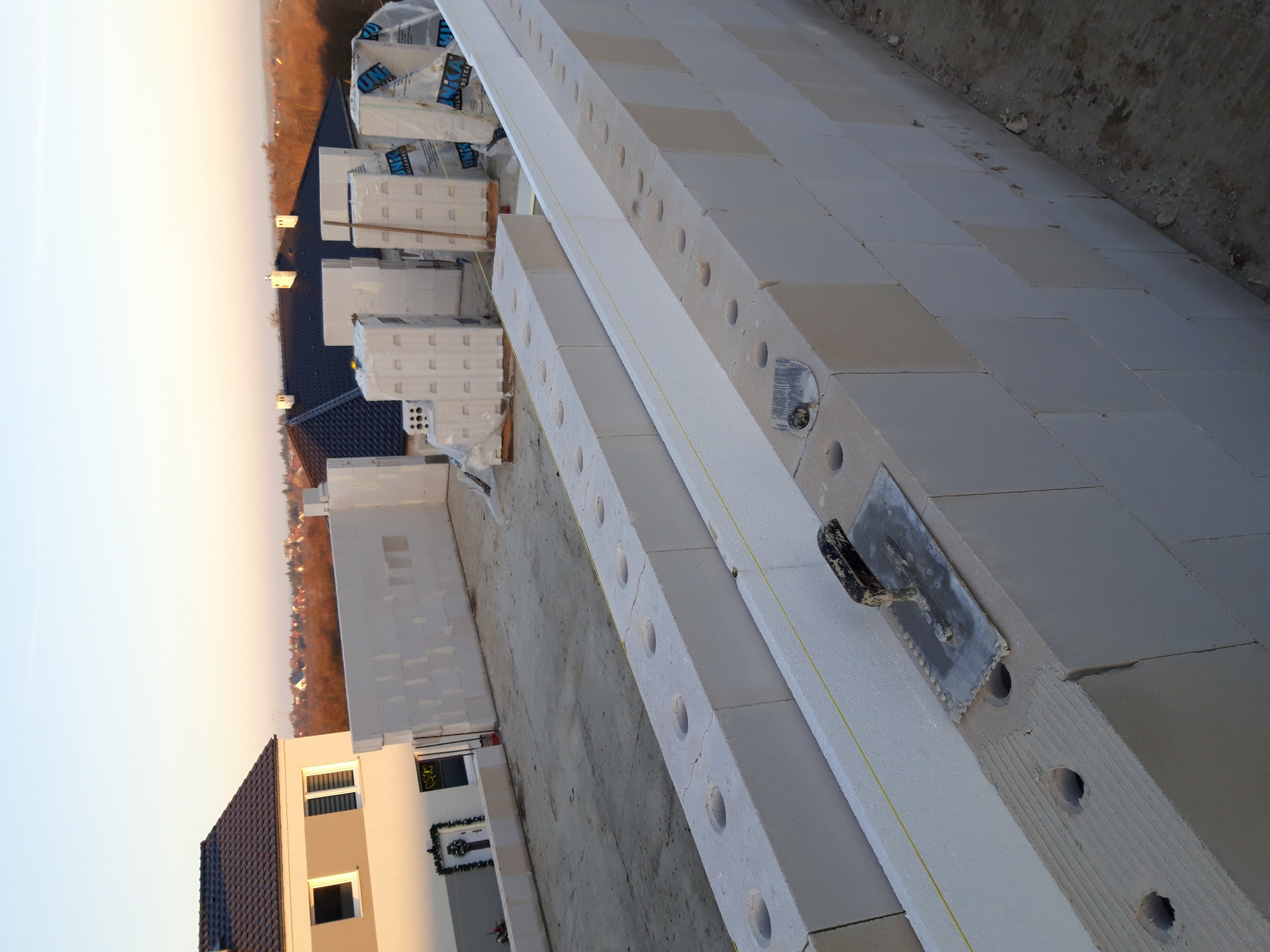 Wznoszenie ściany z betonu komórkowego Ytong, widoczna zaprawa i kielnia murarska, w tle palety z materiałem budowlanym i zabudowa jednorodzinna.