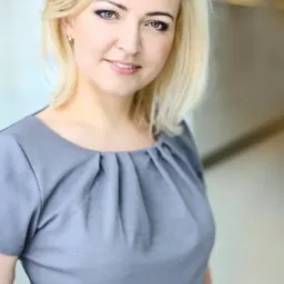 Portret uśmiechniętej blondynki w szarej bluzce, z delikatnym makijażem oczu i subtelną biżuterią w uchu, stojącej na jasnym tle.