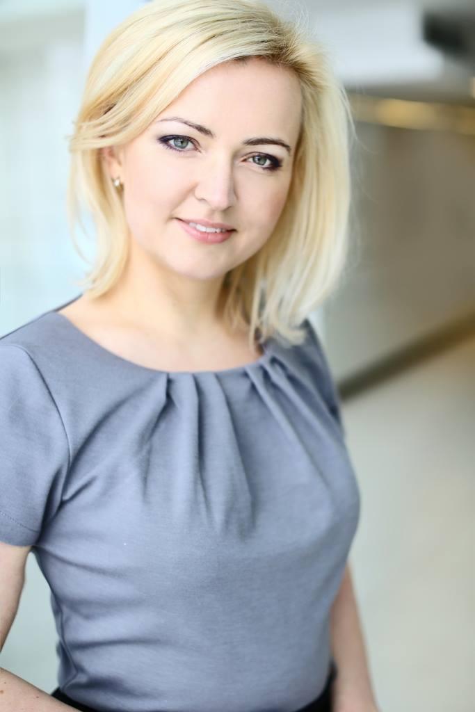 Portret uśmiechniętej blondynki w szarej bluzce, z delikatnym makijażem oczu i subtelną biżuterią w uchu, stojącej na jasnym tle.