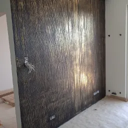 Ściana z dekoracyjną fakturą w odcieniach czerni i złota, widoczne wystające kable elektryczne i gniazdka, podłoga zabezpieczona folią malarską.