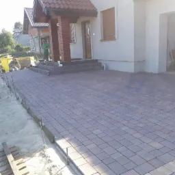 Nowa, brązowo-szara kostka brukowa ułożona przed domem z czerwonym dachem; widoczni dwaj pracownicy podczas układania, jeden pracuje przy piasku, drugi stoi na schodach.