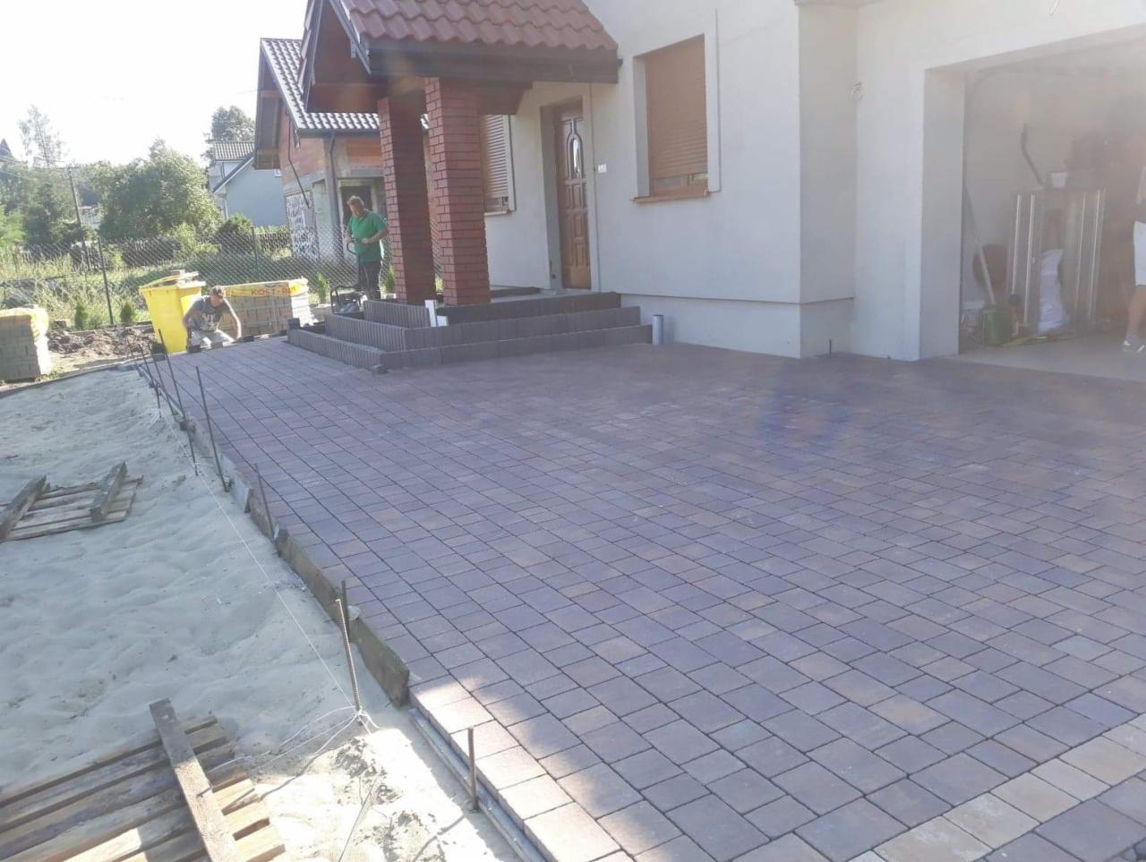 Nowa, brązowo-szara kostka brukowa ułożona przed domem z czerwonym dachem; widoczni dwaj pracownicy podczas układania, jeden pracuje przy piasku, drugi stoi na schodach.