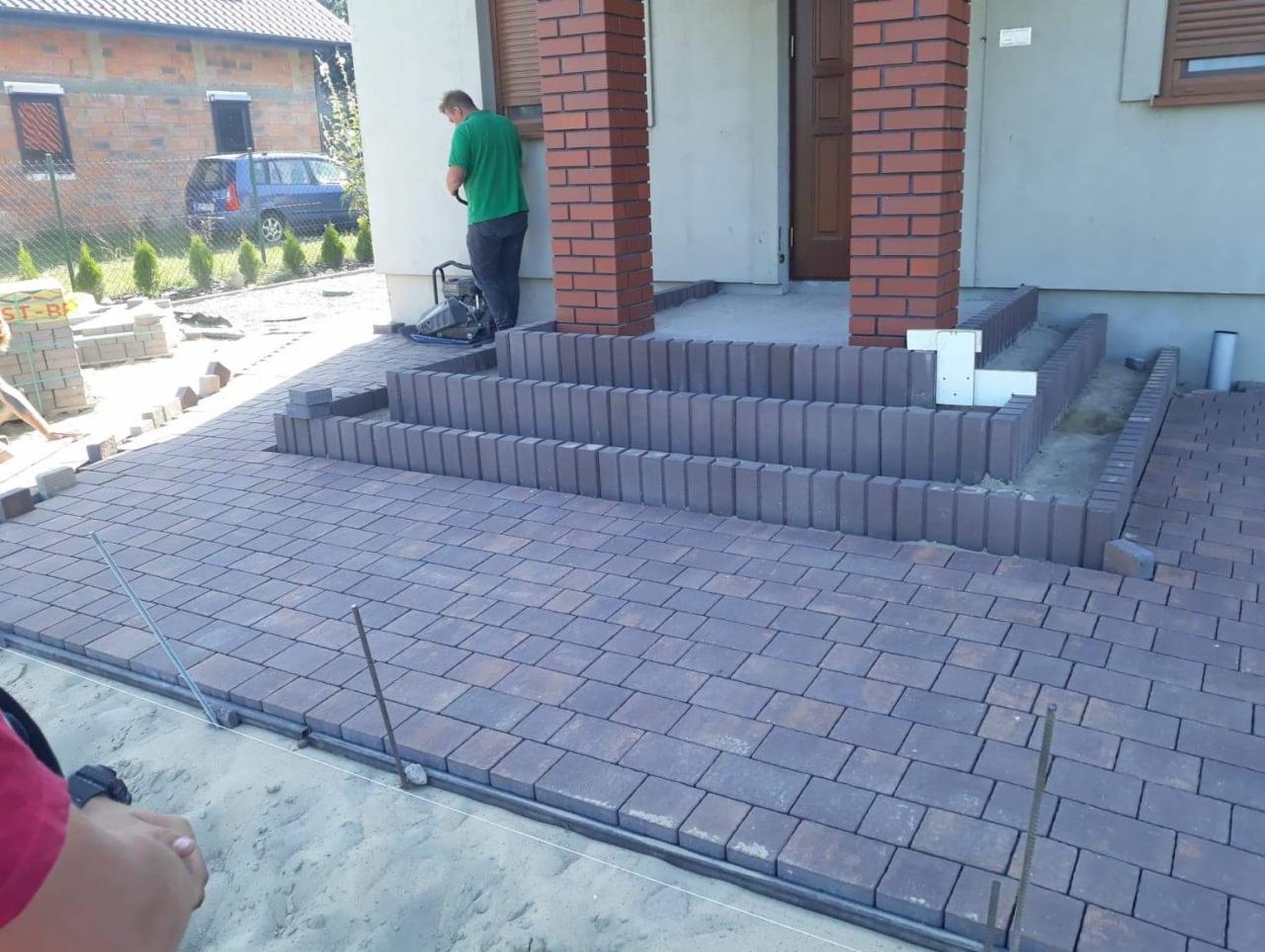Układanie kostki brukowej w odcieniach brązu przed domem, widoczne schody wejściowe i pracownik z zagęszczarką w tle.