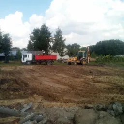 Żółta koparko-ładowarka i czerwona wywrotka na placu budowy z przygotowanym terenem, widoczne ślady opon na ziemi, w tle drzewa i budynki.