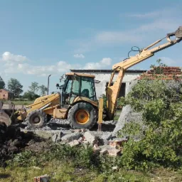Żółta koparko-ładowarka z podniesioną łyżką na tle częściowo zburzonego ceglanego budynku z szarym otynkowaniem, gruz leży na ziemi.
