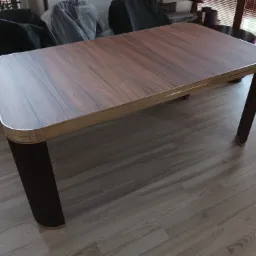 Rozkładany stół z blatem imitującym drewno orzechowe, z zaokrąglonymi rogami i metalową ramą, na ciemnych, żłobkowanych nogach, stojący na panelach podłogowych.