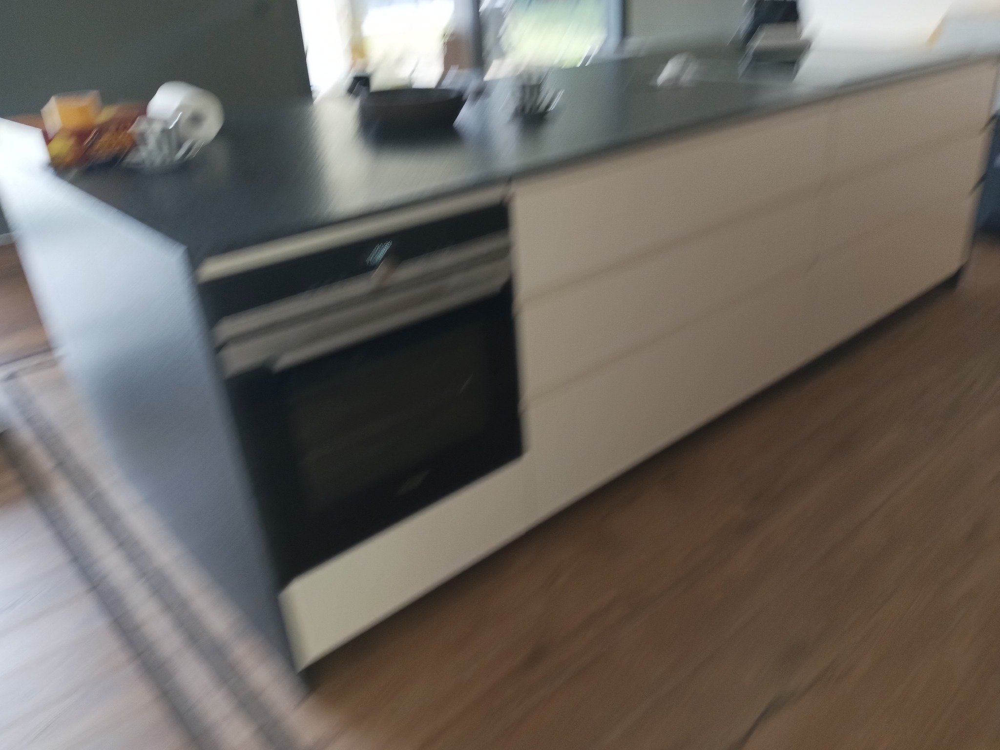 Fragment nowoczesnej wyspy kuchennej z wbudowanym piekarnikiem, blatem w kolorze grafitowym i frontami szafek w odcieniu złamanej bieli, widoczne akcesoria kuchenne na blacie.