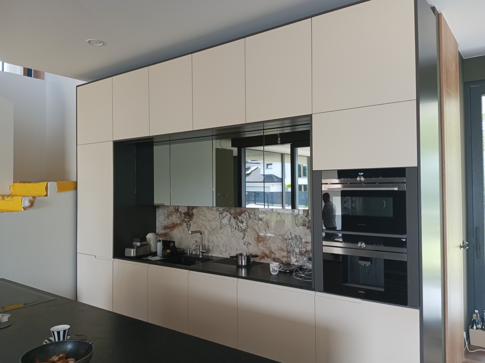 Nowoczesna kuchnia z zabudową w kolorze złamanej bieli, czarnym blatem i marmurowym backsplash. W zabudowie wbudowane piekarniki Siemens. Na blacie widoczny zlew, bateria, czajnik i naczynia.