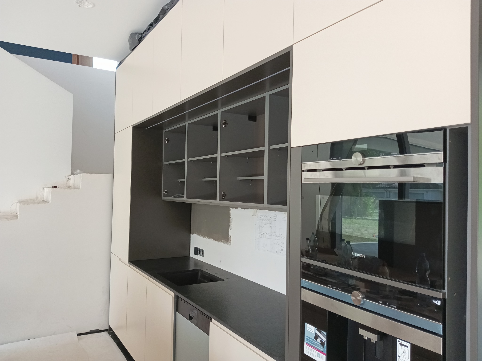 Niedokończona kuchnia z białymi szafkami, czarnym blatem i sprzętem Siemens w nowoczesnym domu z surowymi schodami w tle.