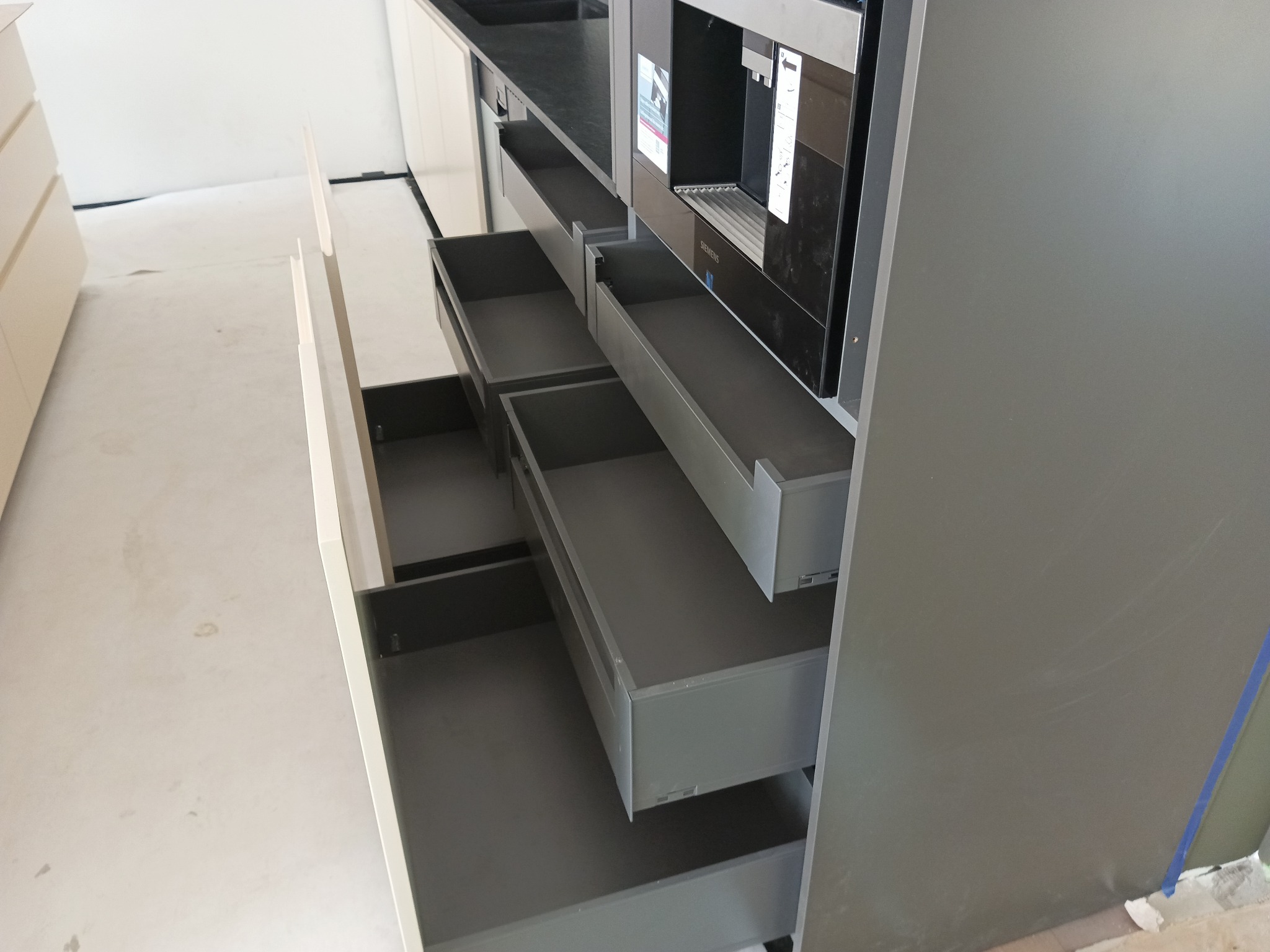 Szare, wysuwane szuflady kuchenne z systemem cichego domykania, zamontowane obok wbudowanego ekspresu do kawy Siemens, widoczne w trakcie montażu na tle surowej podłogi.