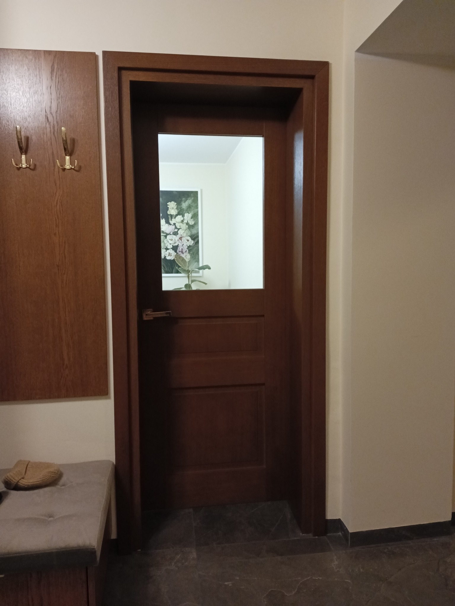 Ciemnobrązowe drzwi wewnętrzne z prostokątnym przeszkleniem w górnej części, widok na jasne wnętrze z obrazem kwiatów, obok wieszak na ubrania i pufa z poduszką.