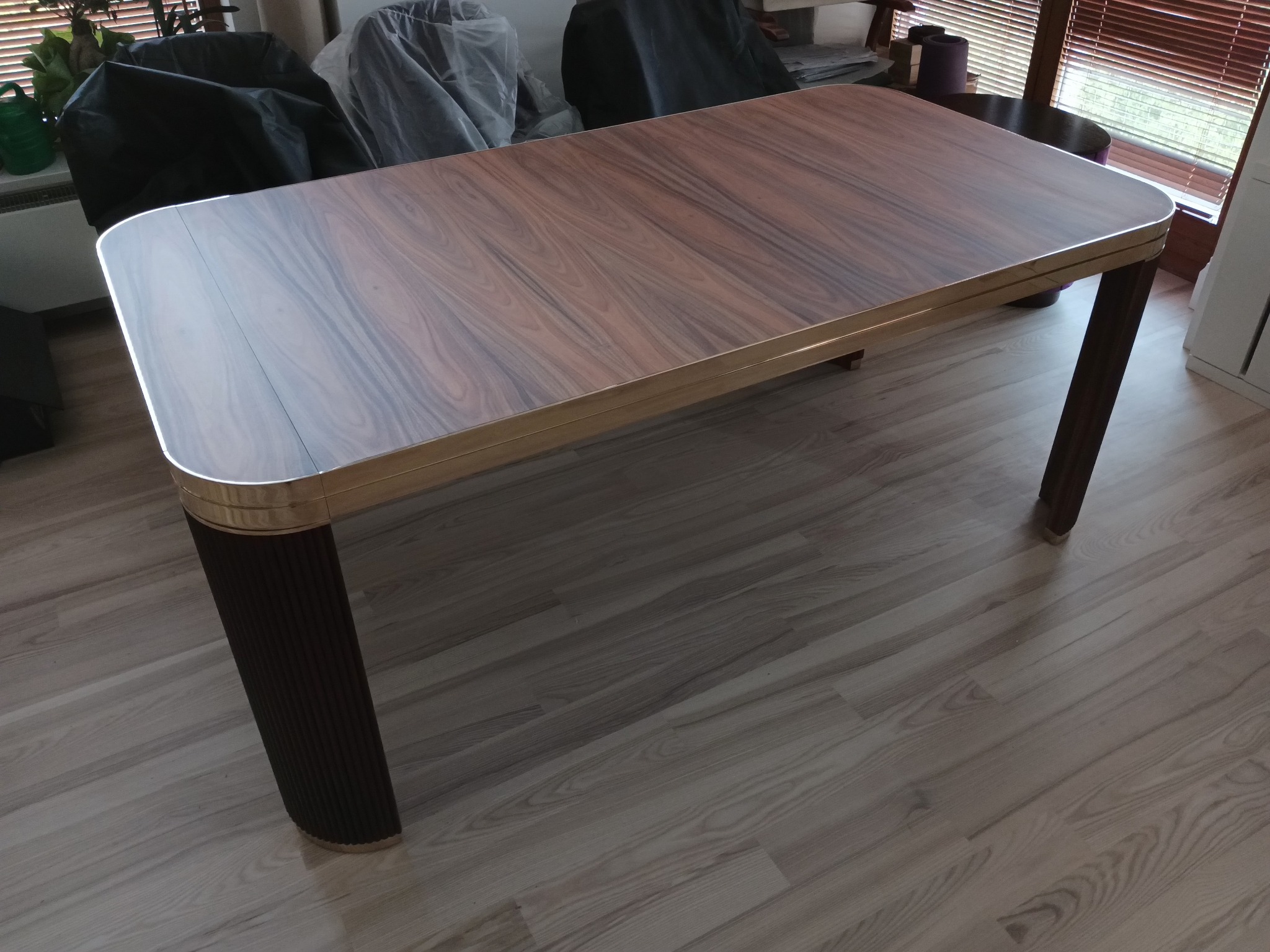 Rozkładany stół z blatem imitującym drewno orzechowe, z zaokrąglonymi rogami i metalową ramą, na ciemnych, żłobkowanych nogach, stojący na panelach podłogowych.