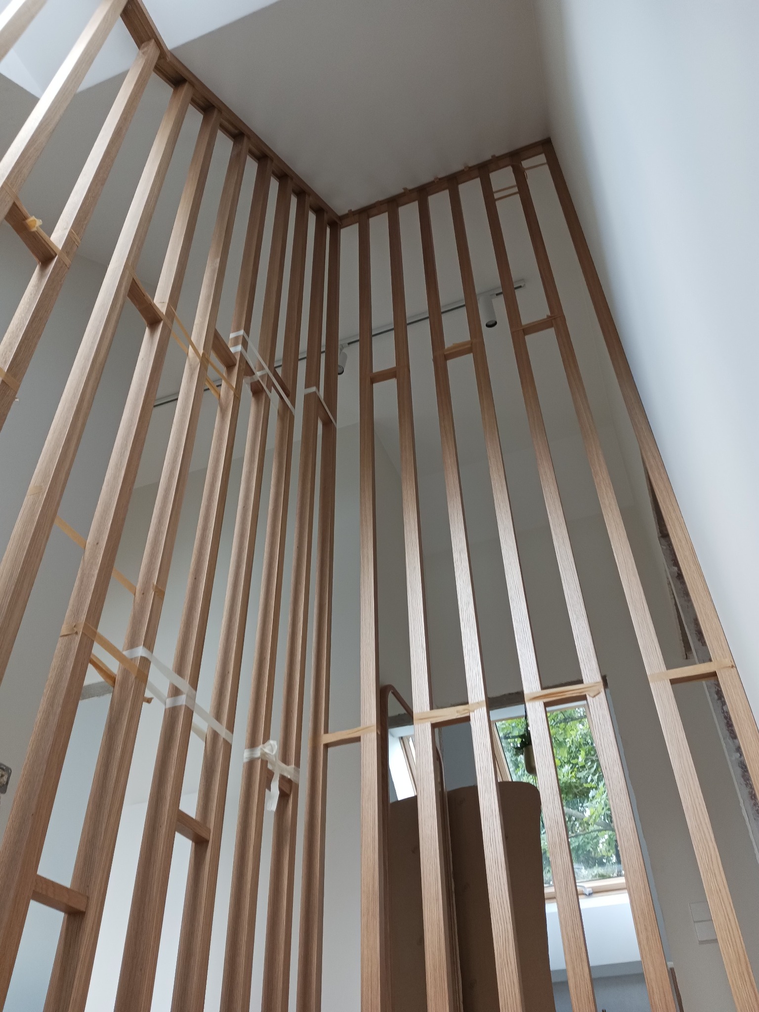 Montaż drewnianej ścianki działowej z pionowych listew, widoczne mocowania taśmami, surowy stan wykończenia wnętrza z oknem w tle.