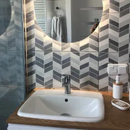 Nowoczesna łazienka z umywalką nablatową na drewnianym blacie, bateria chromowana, geometryczne płytki na ścianie i okrągłe lustro z podświetleniem LED.