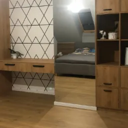 Drewniany regał z biurkiem i szafkami z widocznym odbiciem sypialni w lustrze, na ścianie wzór geometryczny z czarnych linii.