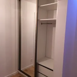 Biała szafa wnękowa z przesuwnymi drzwiami lustrzanymi i widocznymi półkami oraz szufladami, zamontowana w rogu pokoju z drewnianą podłogą.