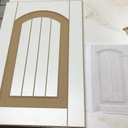 Biały, niedokończony front z płyty MDF z frezowanym łukiem i pionowymi listwami, obok szkic projektu na papierze.