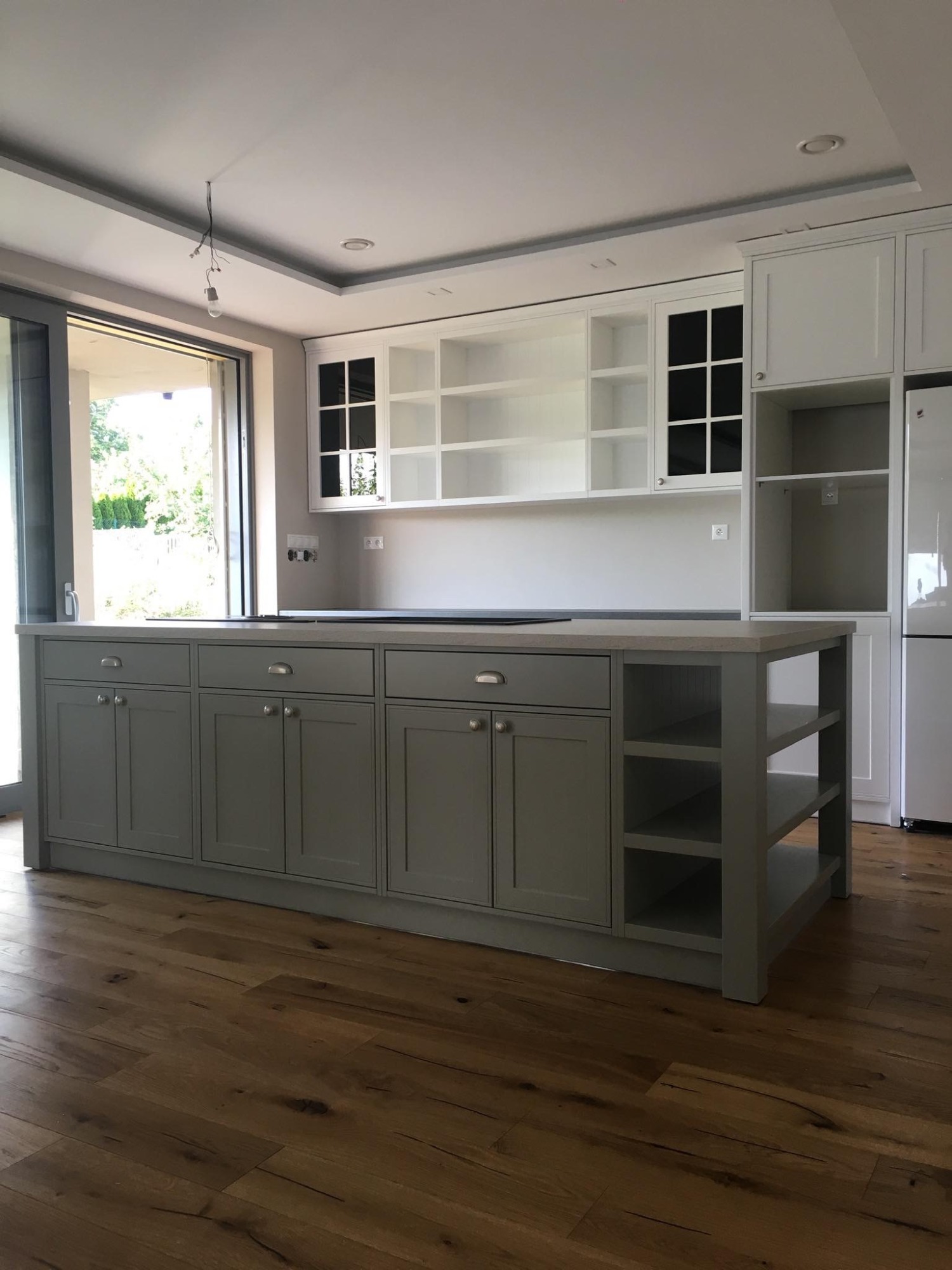 Szara wyspa kuchenna z szafkami i półkami w nowoczesnej kuchni z drewnianą podłogą i białymi szafkami wiszącymi, częściowo przeszklonymi